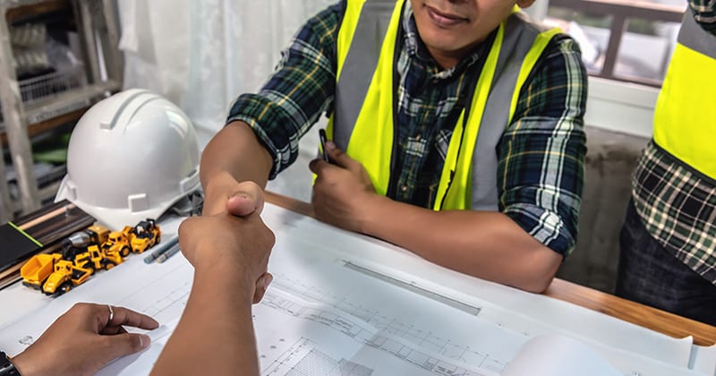 Landscape employee in yellow vest shaking hands with client over printed landscape plans
